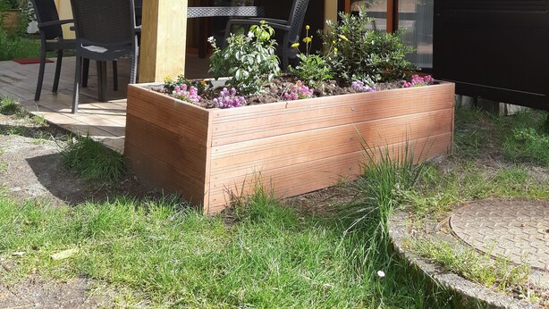 A covered outdoor seating area with a table and chairs, surrounded by a garden bed with various plants and flowers. - Bungalow aux Villages Sous les Pins, à 10 Minutes des Plages (LEON)