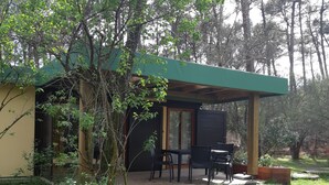 A small, single-story building with a green roof, a covered patio area with a table and chairs, surrounded by trees and greenery. - Bungalow aux Villages Sous les Pins, à 10 Minutes des Plages (LEON)