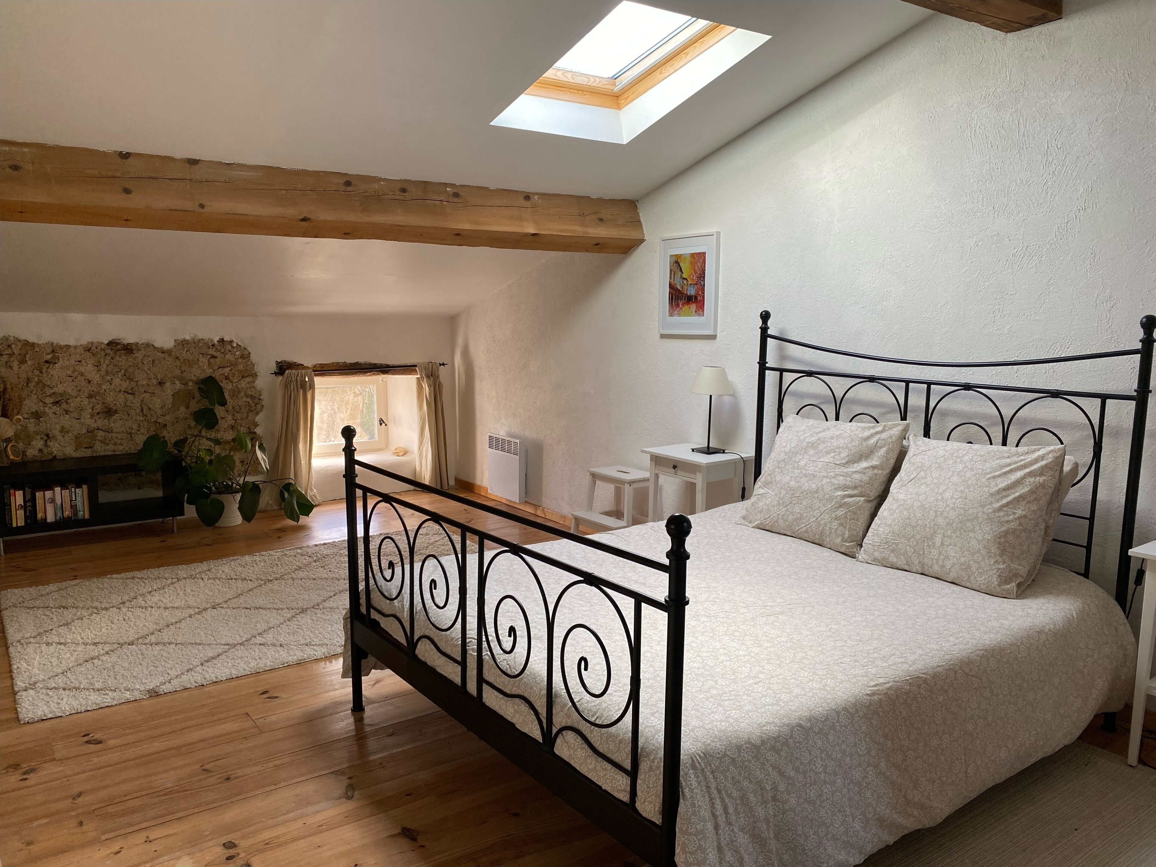 Een slaapkamer met dakraam, een bed met metalen frame, nachtkastje, radiator, stenen muur en een boekenplank.