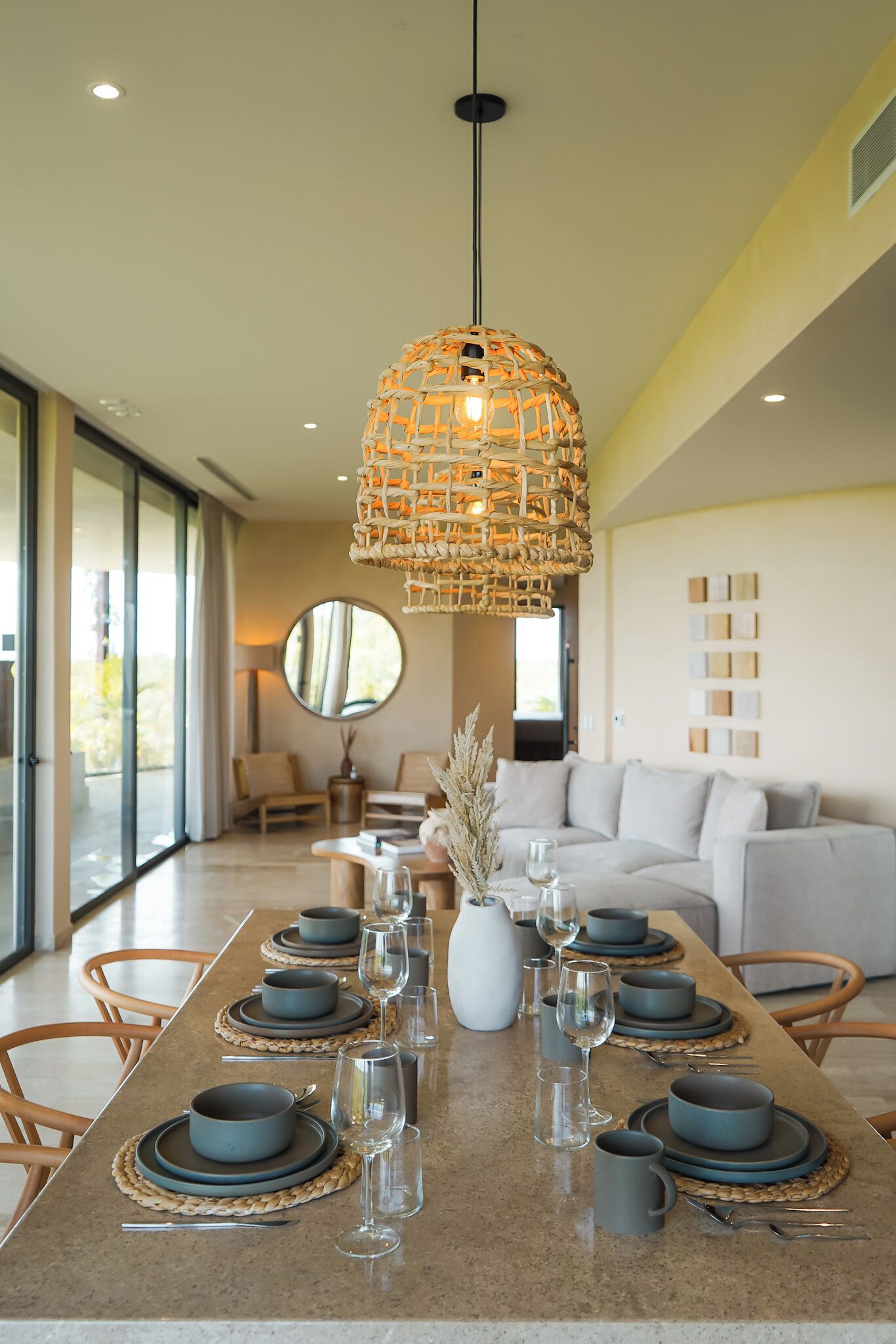 Uma sala de jantar moderna com uma mesa comprida posta para seis pessoas, com jogos americanos trançados e louça de tons escuros. Uma luminária pendente trançada e peculiar paira sobre a mesa, e o ambiente inclui um espelho grande e um sofá confortável.