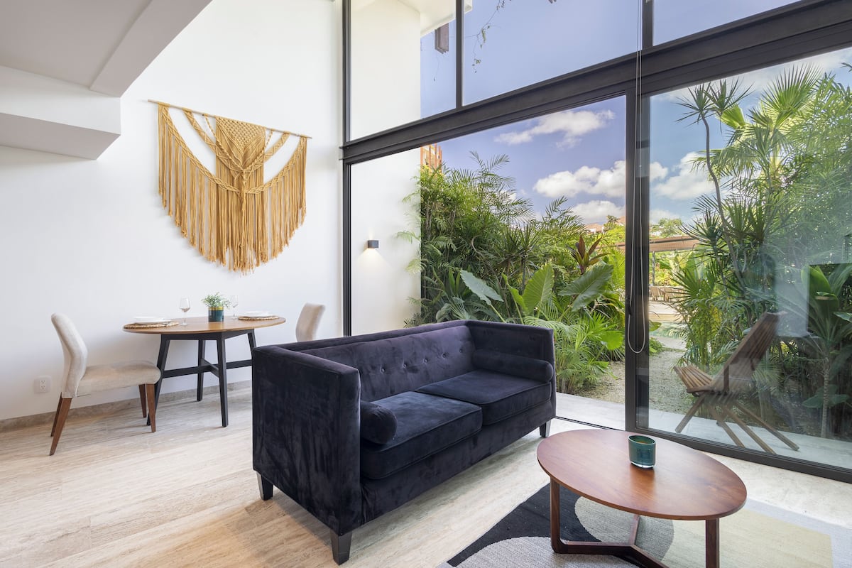 A modern living room with a dark sofa, a round wooden table, and a dining area with a white chair. A macrame wall hanging is visible, and the room has large windows offering a view of greenery outside.