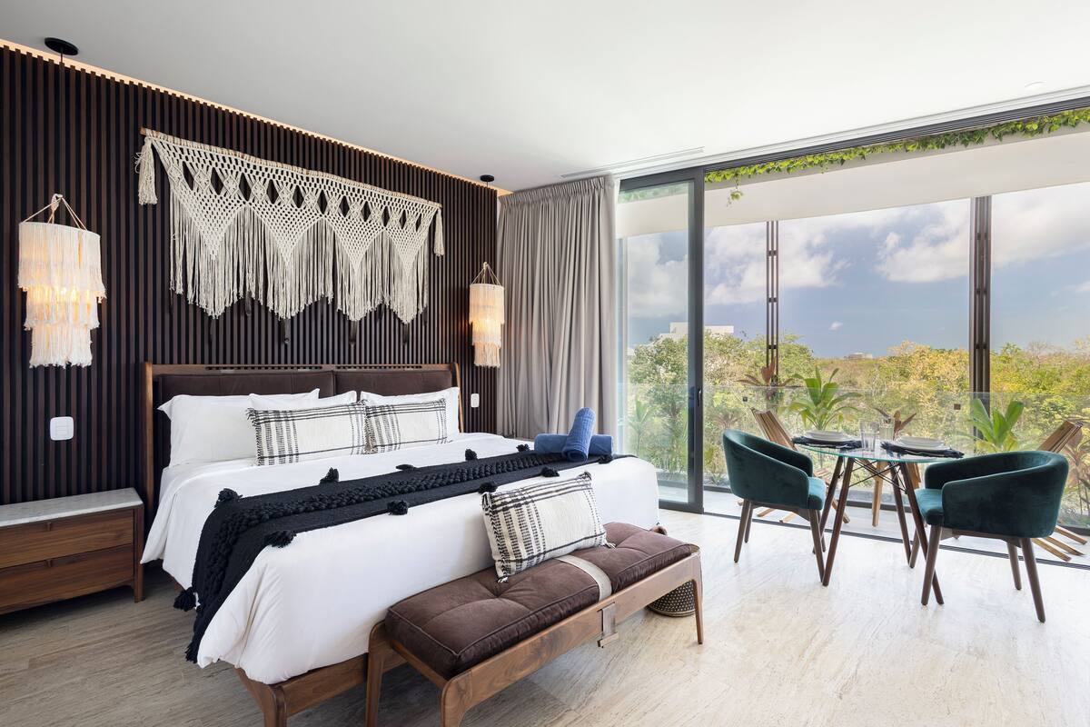 A modern bedroom with a large bed, a bench, a dining table with chairs, and a view of the outdoors.