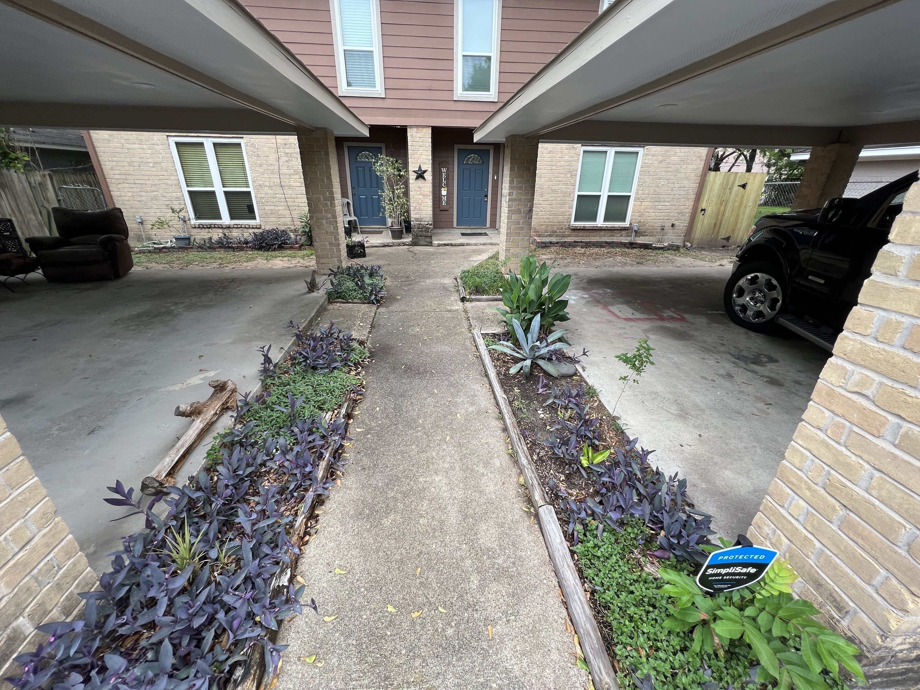 A residential driveway with a car parked on the right, a welcome sign, and a security sign.