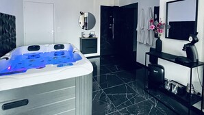 Une salle de bain moderne avec un sol en marbre noir, une baignoire blanche dotée de lumières LED bleues, un miroir rond et un meuble-lavabo noir avec un vasque et un vase de fleurs.