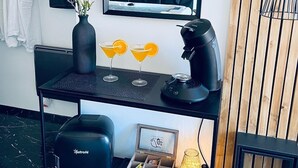 Une salle de bain moderne avec un meuble-lavabo noir, un miroir, un vase avec des fleurs, deux verres à martini, une machine à café, une petite horloge encadrée et une plante décorative.