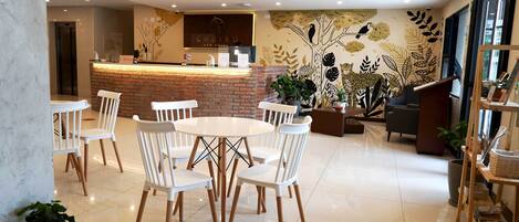 A modern hotel lobby with a round dining table, white chairs, a reception desk, and a mural with nature-themed artwork.