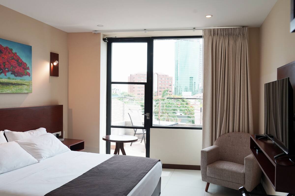 A hotel room with a bed, a desk, a chair, a TV, a painting, and a large window with a city view.