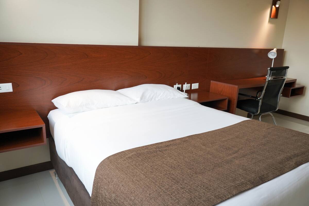 A hotel room with a wooden headboard, a bed with white linens, a brown throw blanket, a desk with a chair, and a wall-mounted lamp.