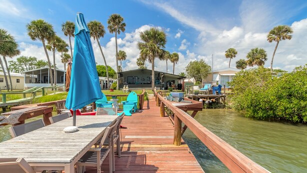A wooden deck with tables and chairs, overlooking a body of water with palm trees and a building in the background.