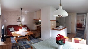A cozy living room with a red sofa, a wooden coffee table, and a dining table with a vase of flowers. There is a kitchen area in the background with cabinets and a hanging light fixture.