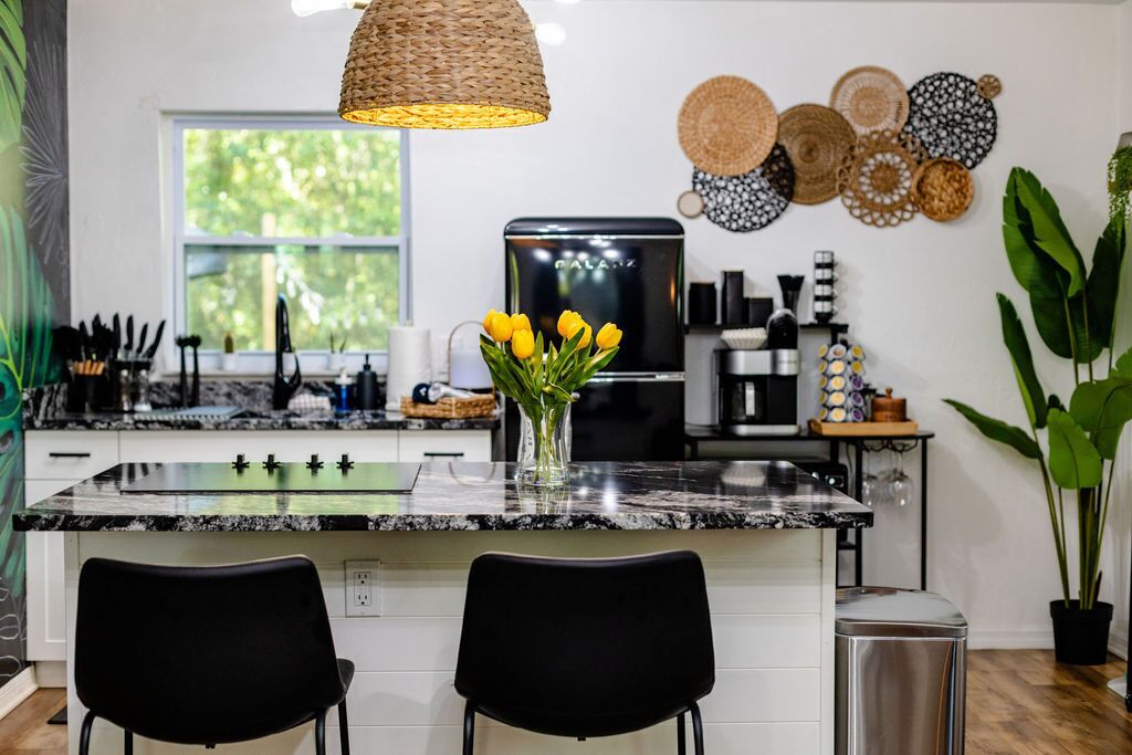 A modern kitchen with a granite countertop, black chairs, a woven pendant light, and a vase with yellow tulips.