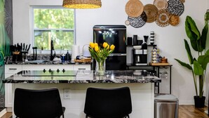 Eine moderne Küche mit einer Granit-Arbeitsplatte, schwarzen Stühlen, einer geflochtenen Pendelleuchte und einer Vase mit gelben Tulpen.