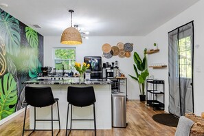 A modern kitchen with a central island, black bar stools, a hanging woven light fixture, and a large green plant.