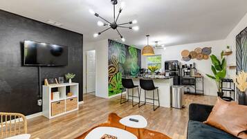 A modern living room with a flat-screen TV, a dining area with a bar, and a kitchen with a coffee machine.