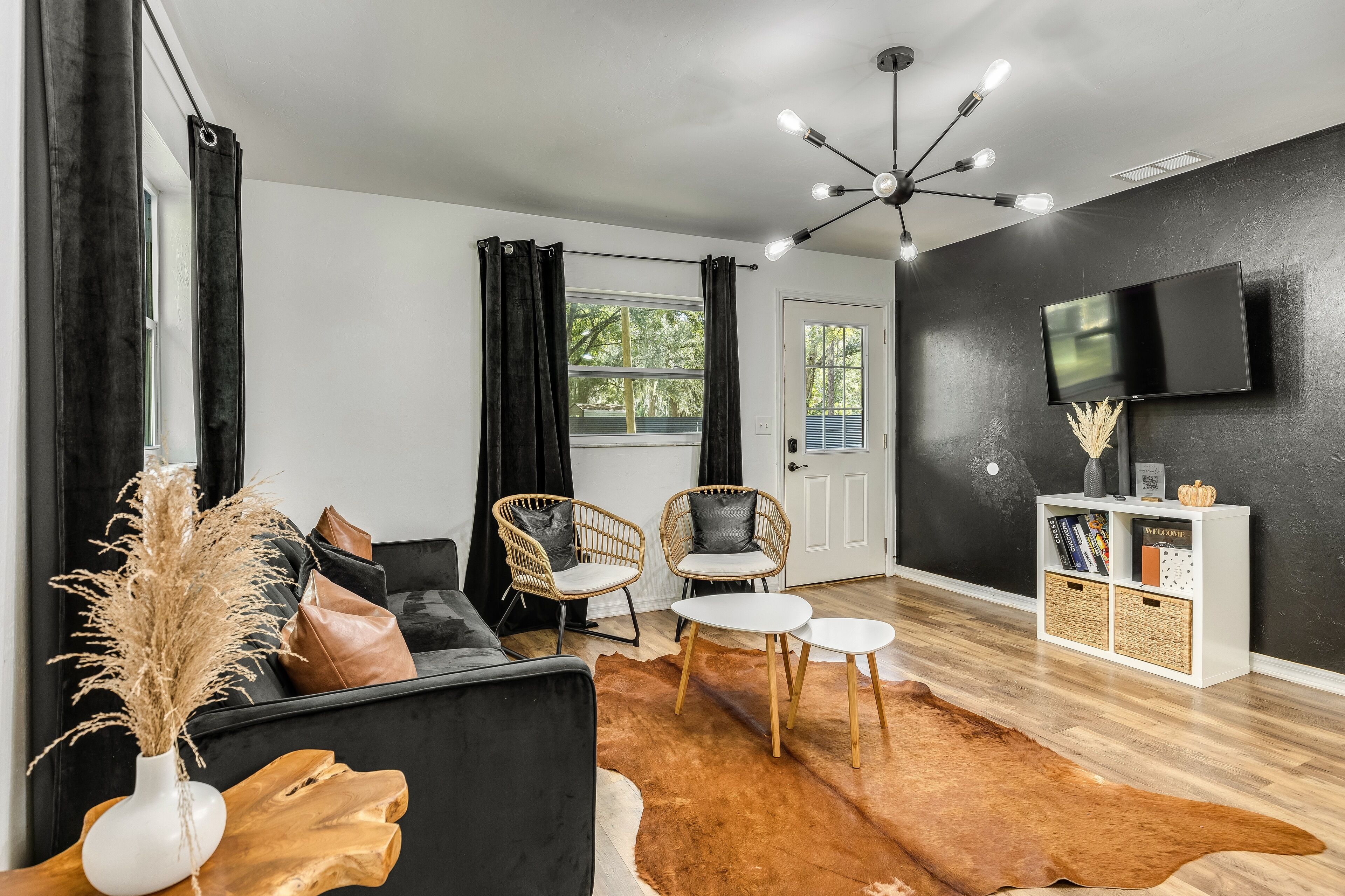 A modern living room with a black sofa, two wicker chairs, a coffee table, a wooden side table, a TV on the wall, and a bookshelf.