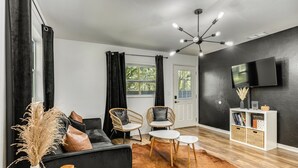 Ein modernes Wohnzimmer mit einer schwarzen Couch, zwei Korbstühlen, einem Couchtisch, einem Holznachttisch, einem Fernseher an der Wand und einem Bücherregal.