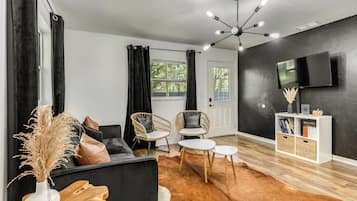 A modern living room with a black sofa, two wicker chairs, a coffee table, a wooden side table, a TV on the wall, and a bookshelf.