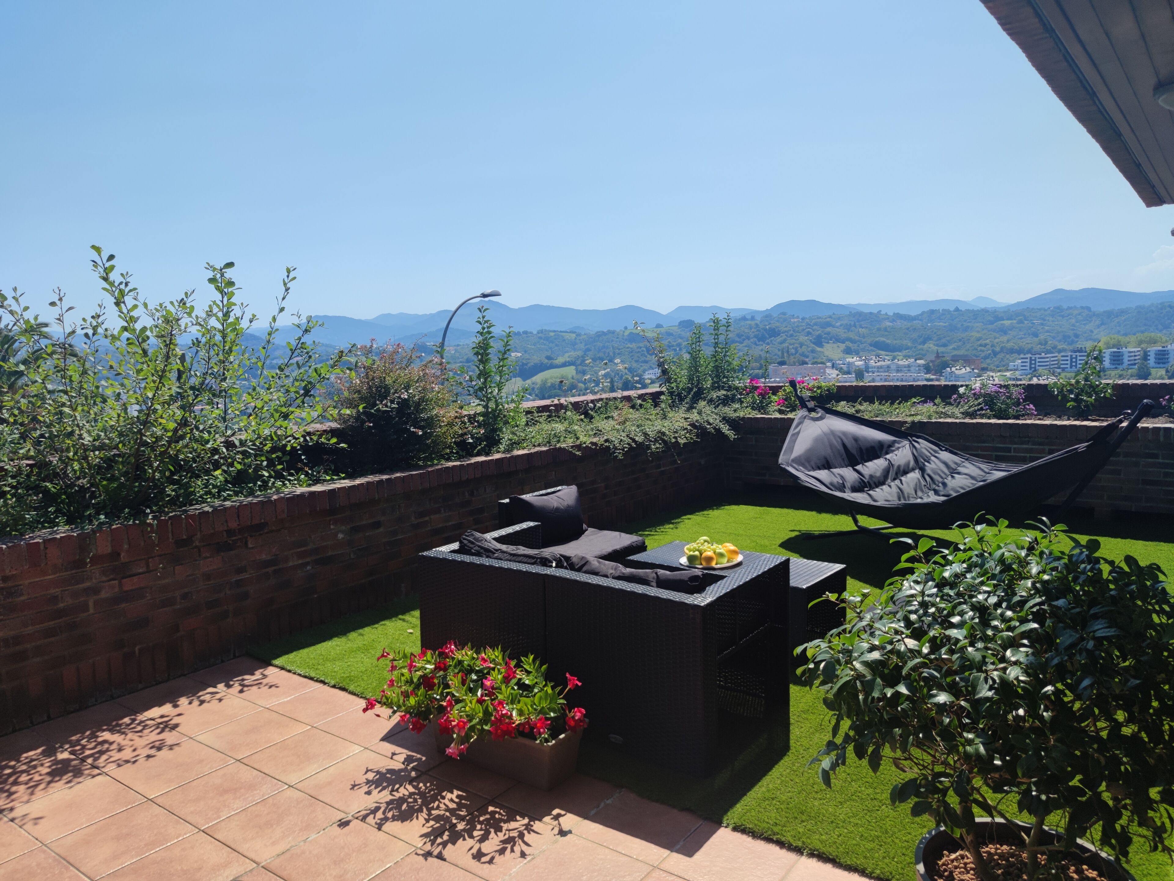 Un patio con un sofá exterior negro, una hamaca y una pequeña mesa con fruta. Hay plantas en macetas y se ve una vista de montañas a lo lejos.