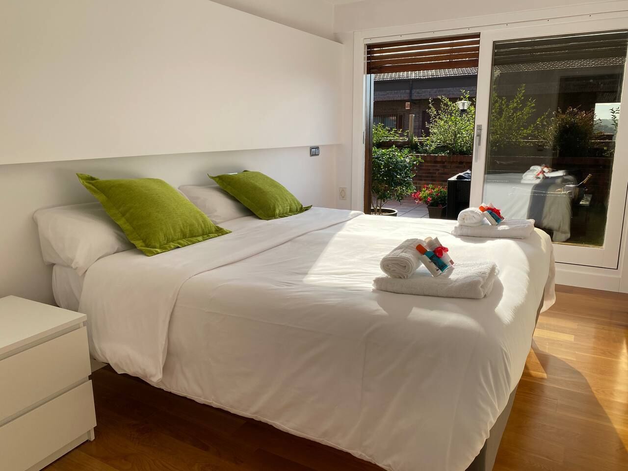 A modern bedroom with a large bed, white bedding, and green pillows. There are towels on the bed and a white nightstand to the left.