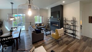 Een moderne woonkamer met een grote glazen salontafel, een bank, een gele fauteuil, een open haard en een aan de muur bevestigde televisie.