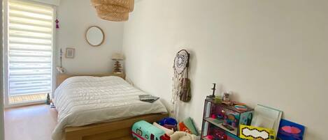 A bedroom with a bed, a hanging light fixture, a window with blinds, a mirror, and a toy storage unit.
