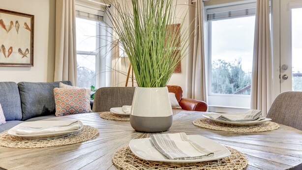 Un coin repas avec une table en bois dressée pour quatre personnes, un vase contenant des plantes vertes et une vue sur l’extérieur grâce à de grandes fenêtres.