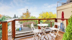 Une terrasse en bois avec une table blanche et deux chaises, un parasol rouge, et une vue sur un jardin avec des arbres et un abri de jardin.