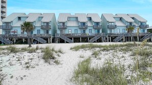 Ein Strandhotel mit mehreren Gebäuden, die durch Laubengänge verbunden sind, mit Balkonen und einer Sandstrandfläche.