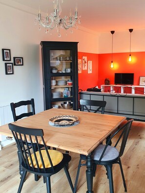 Een eetruimte met een houten tafel en zwarte stoelen, een kroonluchter, en een keuken met een rode muur en hangende lichtbollen.