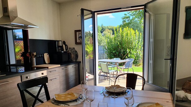 A dining area with a table set for four, a kitchen in the background, and a view of a garden through a glass door. - The calm, the charm, the Maison du Menez: "the place to be" (La Forêt-Fouesnant)