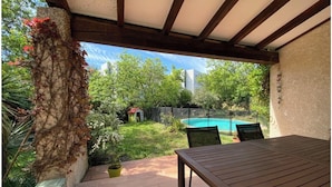 Une terrasse couverte avec une table et des chaises en bois, une piscine et une végétation luxuriante.