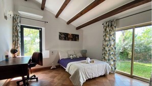 Une chambre à coucher avec un lit, un bureau et une chaise. Il y a une fenêtre avec des rideaux, un tableau au mur et une vue sur l’extérieur.