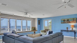 A living room with a large sectional sofa, a ceiling fan, and large windows overlooking a body of water.