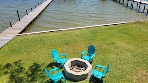 A lakeside deck with a fire pit and Adirondack chairs, a dock extending into the water, and a covered area at the end of the dock.