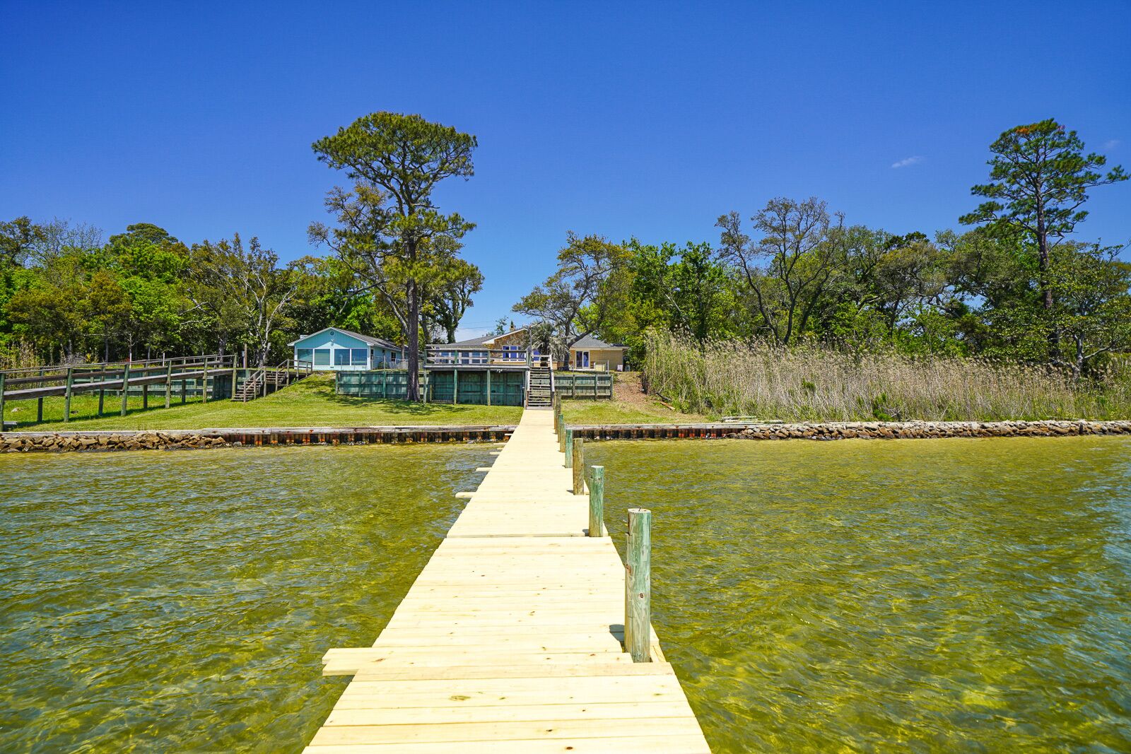 A wooden pier leading to a dock, surrounded by greenery and clear water.
