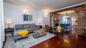 Ein modernes Wohnzimmer mit einem grauen Sofa, einem Couchtisch und einem gemusterten Teppich. Es gibt einen Essbereich mit einer Ziegelwand und einer Chandelier.