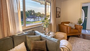 A living room with a sofa, a coffee table, and a chair. There is a window with curtains, a plant on the table, and a door in the background. - Green Door Getaway — family-friendly 4BR, 10 minutes to New River Gorge (Oak Hill)