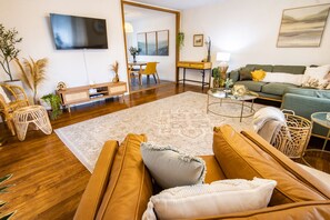 A modern living room with a wooden floor, a large flat-screen TV mounted on the wall, a green sofa, a yellow armchair, and a coffee table.