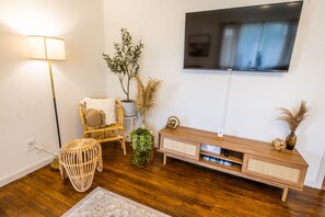 A modern living room with a wooden floor, a mounted TV, a wicker chair, a small side table, and a console table with decorative items.
