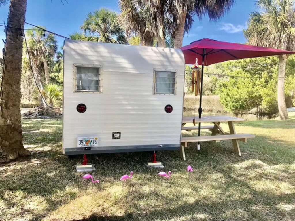 A white RV with a small window and a license plate, parked on grass with a picnic table and umbrella nearby.