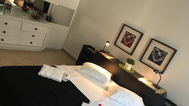Een net opgemaakt bed met witte lakens, een nachtkastje met een lamp, en twee ingelijste kunstwerken aan de muur.