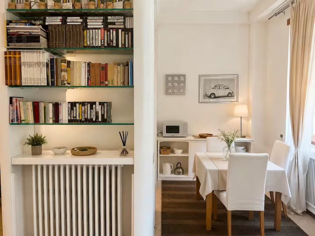 Una habitación con una estantería, un comedor con mesa y sillas, un microondas y un cuadro en la pared.