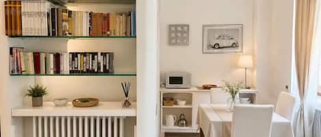 Una habitación con una estantería, un comedor con mesa y sillas, un microondas y un cuadro en la pared.
