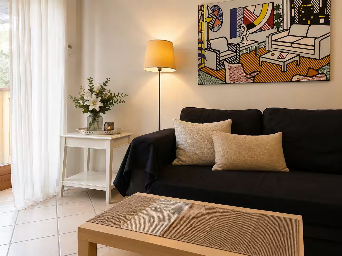 Un soggiorno con un divano nero, un piccolo tavolino con una lampada e un quadro appeso al muro.