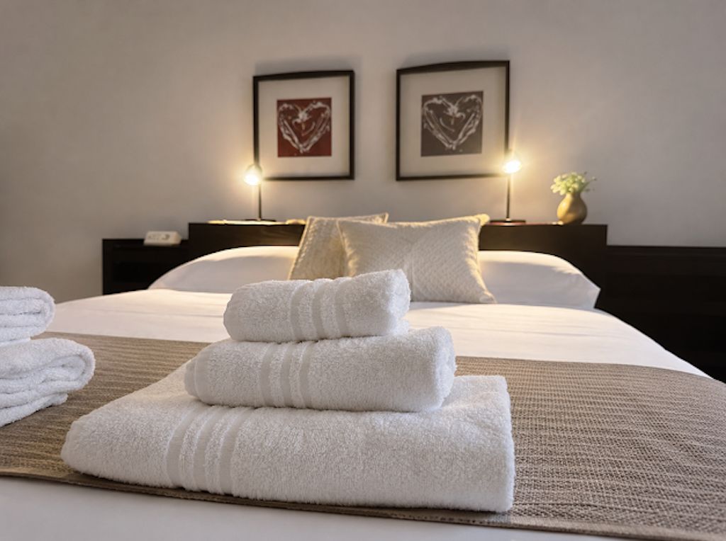 A hotel room with a neatly made bed, two framed heart-shaped pictures on the wall, and a bedside table with a lamp and a vase.
