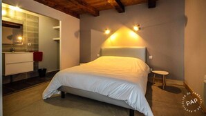 A bedroom with a large bed, a wooden ceiling, a small stool, and a bathroom with a mirror and white cabinet. - Mas des Pêcheurs (Le Ségur)