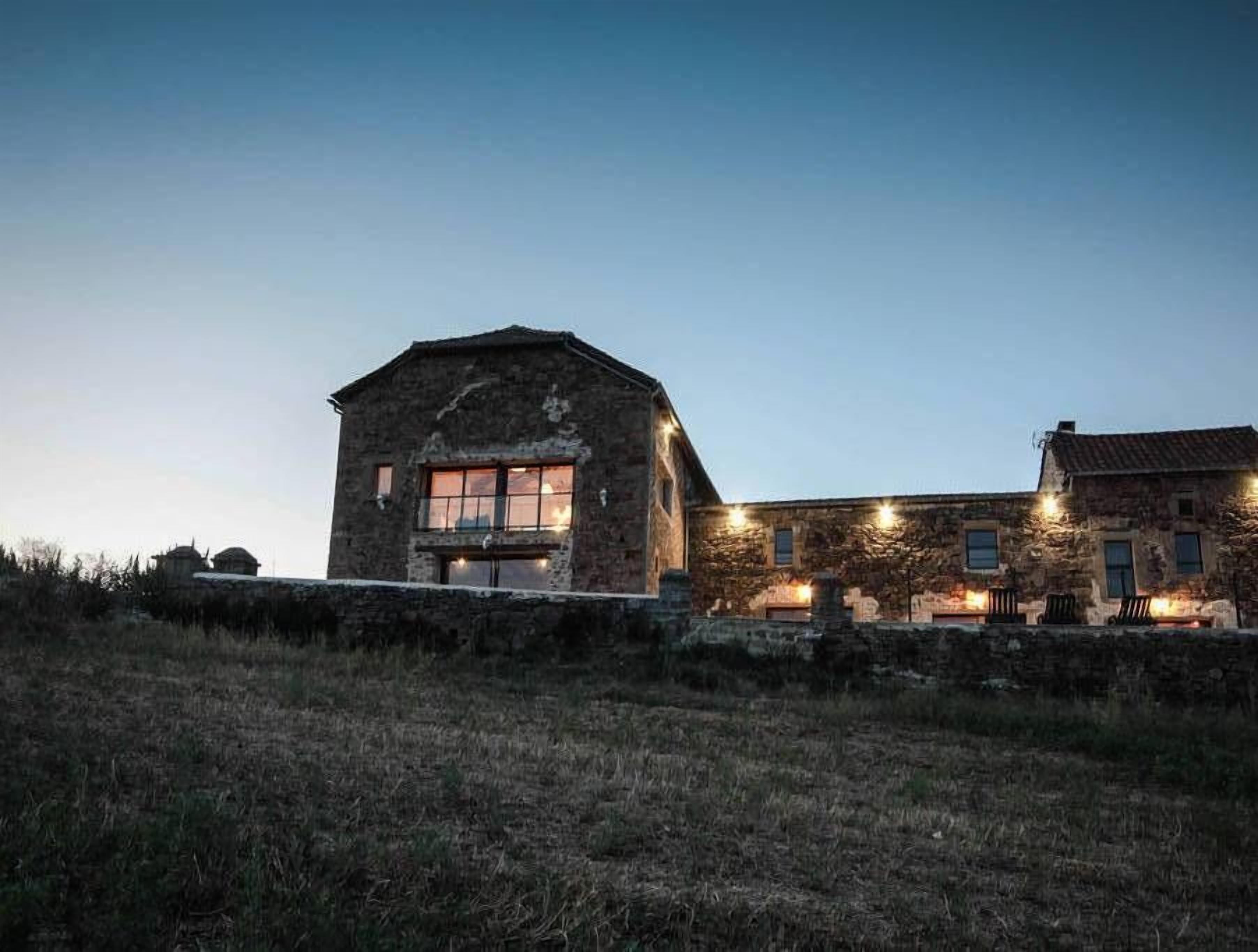 A stone building with a large window and a smaller window on the side, surrounded by a stone wall and a grassy area.