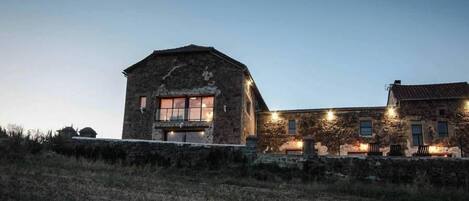 A stone building with a large window and a smaller window on the side, surrounded by a stone wall and a grassy area.