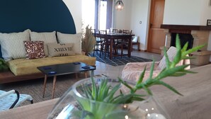 Olohuoneessa on sohva, sohvapöytä, ruokapöytä ja tuoleja. Etualalla on takka ja kasvi.