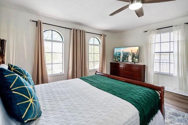 A bedroom with a bed, a dresser, a television, and windows with curtains.